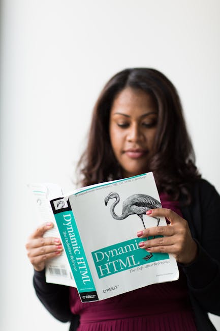A woman with shoulder-length dark hair, wearing a black blazer and maroon top, is reading a book titled 'Dynamic HTML' published by O'Reilly. She is holding the book with both hands, focusing her gaze